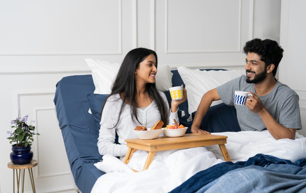 A couple sits upright in an adjustable bed, each holding a mug. The woman has a breakfast tray on her lap, and the elevated bed supports them in a comfortable sitting position.